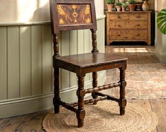 May include: A dark brown wooden chair with a square seat and a backrest featuring a decorative panel with gold and brown painted floral patterns. The chair has four legs connected by horizontal stretchers, all with a turned, rounded design.