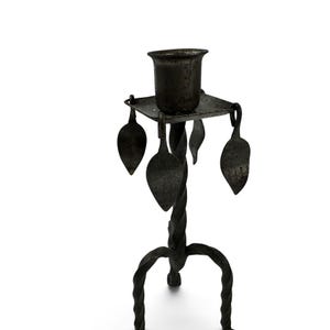 May include: A dark gray metal candle holder featuring a central twisted stem, a square top platform, and three curved legs. Four flat, leaf-shaped metal ornaments hang from the corners of the square platform, surrounding the central candle cup.