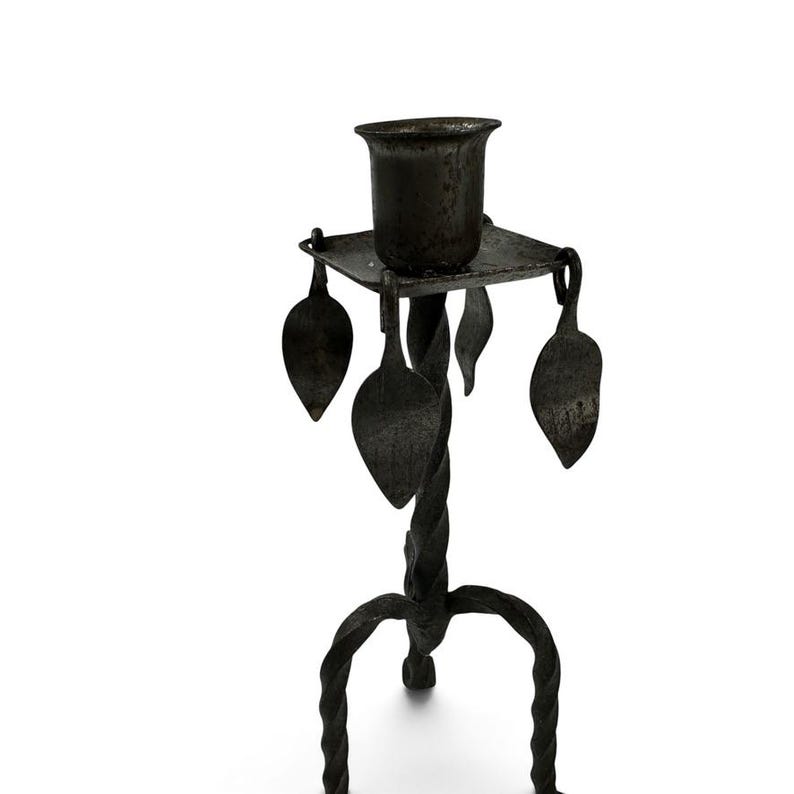 May include: A dark gray metal candle holder featuring a central twisted stem, a square top platform, and three curved legs. Four flat, leaf-shaped metal ornaments hang from the corners of the square platform, surrounding the central candle cup.