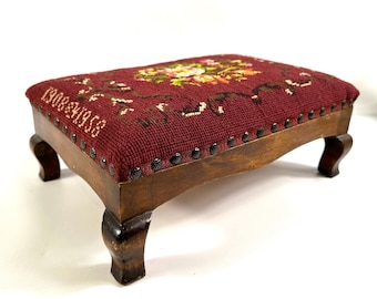 May include: A rectangular footstool with a dark wooden frame and four curved legs. The top is upholstered in a deep burgundy needlepoint fabric featuring a floral design and the embroidered text 1908-1958, secured with black metal nailhead trim.