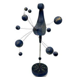 May include: A blue wooden sculpture featuring a central vertical post on a circular base, topped with a stylized bird-like form. Thin metal wires extend outward, each ending in a small blue sphere with white painted details and worn edges.