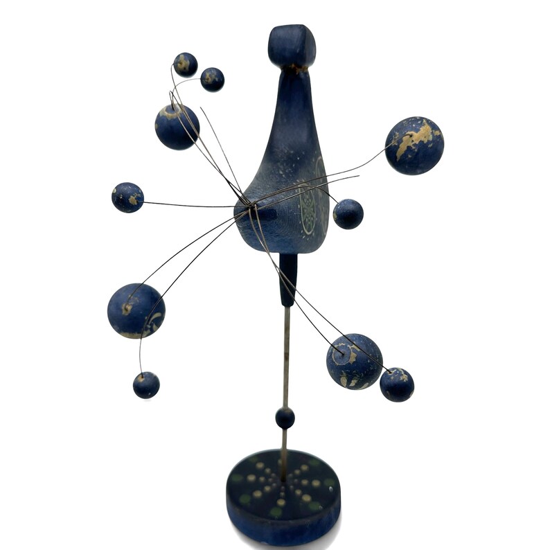 May include: A blue wooden sculpture featuring a central vertical post on a circular base, topped with a stylized bird-like form. Thin metal wires extend outward, each ending in a small blue sphere with white painted details and worn edges.
