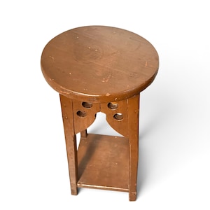 May include: A brown wooden plant stand featuring a circular top surface, four straight legs, and a lower square shelf. The upper section of the stand includes decorative cutouts with circular holes near the top of each leg.