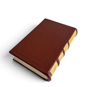 Puede incluir: Un objeto decorativo de cer&aacute;mica rectangular con forma de libro cerrado con una cubierta de color marr&oacute;n oscuro lisa y brillante. El lomo presenta tres bandas en relieve con detalles amarillos y un peque&ntilde;o motivo floral blanco, los bordes son color crema.