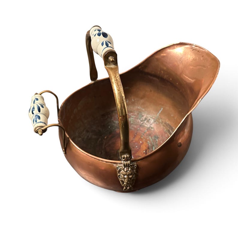 Pu&ograve; includere: Un secchio per carbone in rame con un ampio beccuccio e un manico in ottone caratterizzato da un supporto a testa di leone. Il manico e l'impugnatura laterale presentano inserti in ceramica bianca decorati con motivi floreali blu.
