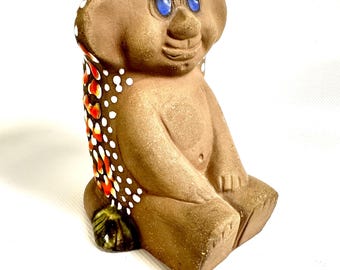 May include: A brown ceramic troll figurine with large ears and bright blue eyes. The figure features white polka dots on the head and a side panel decorated with a pattern of orange, red, and white dots. It sits in a seated position with feet forward.