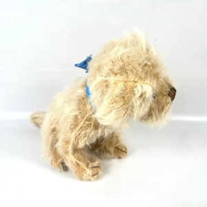 以下が含まれることがあります： ベージュの毛並みと青い首輪が特徴の、ヴィンテージの犬のぬいぐるみ。犬は座っており、頭を横に向け、茶色の鼻と垂れ耳を見せています。おもちゃはクラシックで愛らしい外観をしています。