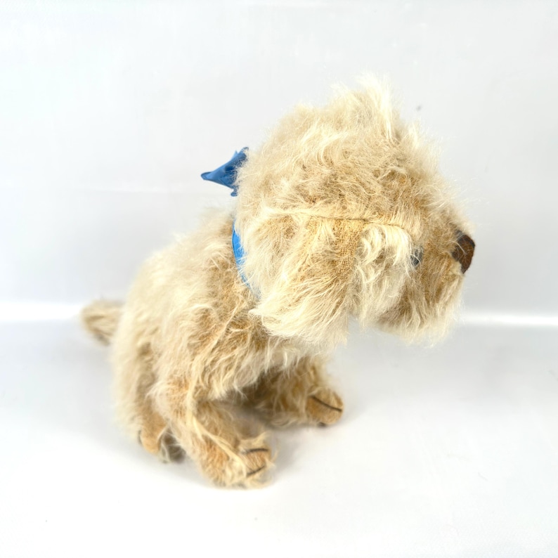 以下が含まれることがあります： ベージュの毛並みと青い首輪が特徴の、ヴィンテージの犬のぬいぐるみ。犬は座っており、頭を横に向け、茶色の鼻と垂れ耳を見せています。おもちゃはクラシックで愛らしい外観をしています。