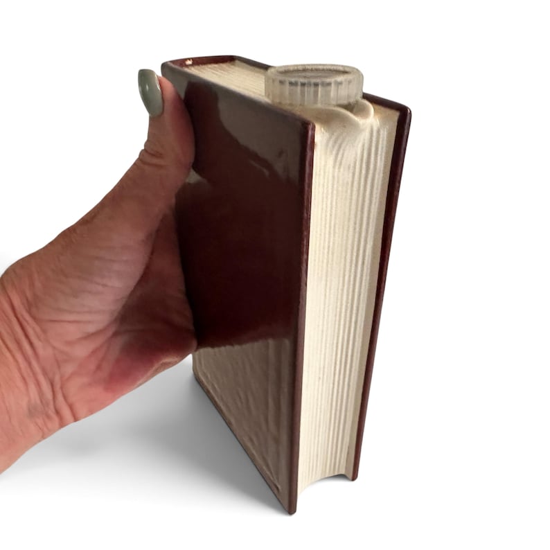 Puede incluir: Una petaca de cer&aacute;mica marr&oacute;n con forma de libro, cuerpo rectangular y acabado brillante. El lado que representa las p&aacute;ginas tiene una textura de estr&iacute;as verticales en color blanco roto. Incluye un tap&oacute;n de rosca de pl&aacute;stico transparente.