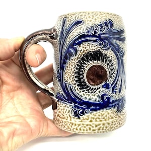 Puede incluir: Una jarra de cerveza de cer&aacute;mica con un acabado texturizado en beige y bronceado, decorada con patrones de volutas florales en azul cobalto y un medall&oacute;n circular marr&oacute;n en el centro. La jarra tiene un asa curva con un acento marr&oacute;n oscuro.