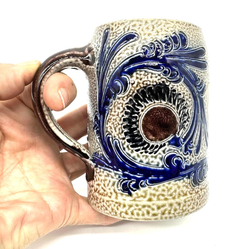 Puede incluir: Una jarra de cerveza de cer&aacute;mica con un acabado texturizado en beige y bronceado, decorada con patrones de volutas florales en azul cobalto y un medall&oacute;n circular marr&oacute;n en el centro. La jarra tiene un asa curva con un acento marr&oacute;n oscuro.