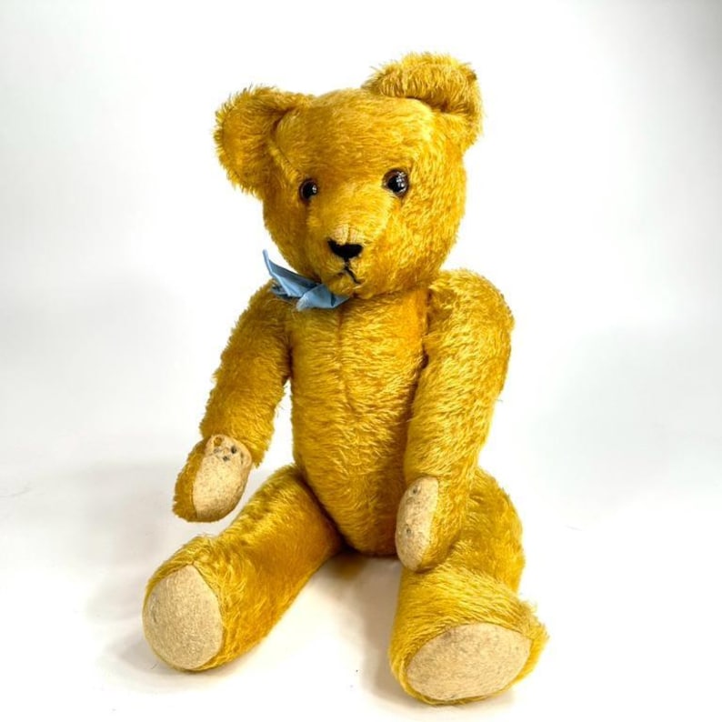 May include: A vintage golden-yellow teddy bear with a light blue bow tie. The bear has black eyes, a black nose, and beige paw pads. It is sitting upright with its arms and legs extended.