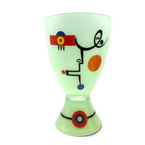K&ouml;nnte beinhalten: Ein blassgr&uuml;ner Glasbecher mit einer Reihe abstrakter geometrischer Formen und Linien in Schwarz, Rot, Blau und Orange. Das Design umfasst einen roten rechteckigen Block mit einem gelben und blauen Kreis, einen gro&szlig;en orangefarbenen Kreis und ein Basismotiv.