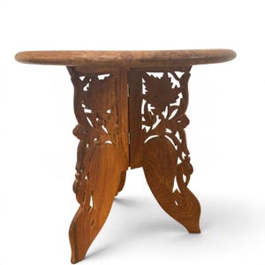 May include: A round wooden table with a smooth top and a folding base. The base consists of two interlocking panels featuring intricate, hand-carved floral and leaf patterns. The wood has a natural medium-brown grain and finish.