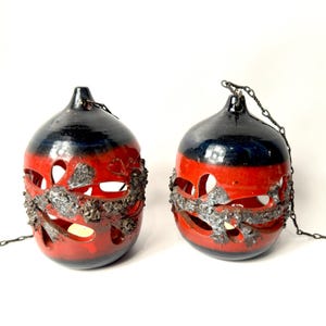 May include: Two spherical ceramic hanging lanterns featuring a glossy red and black glaze. The lanterns have cut-out petal shapes and a textured, rough gray band encircling the middle. Each piece includes a metal chain for suspension.
