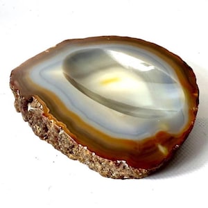 Op de afbeelding: Een kom van gepolijste agaatsteen met een glad, concaaf midden met concentrische banden van wit, grijs en amber. De buitenrand van de steen blijft in zijn natuurlijke, ruwe en getextureerde staat en vertoont een diepbruine en aardse kleur.