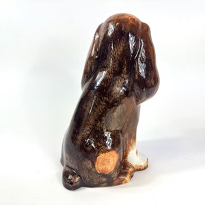 Peut inclure: Figurine en c&eacute;ramique repr&eacute;sentant un chien, vue de dos. Le chien est principalement marron avec des marques marron fonc&eacute; et beige. La figurine a une finition brillante et est assise.