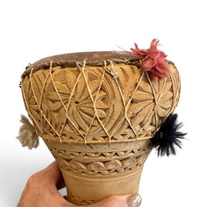 May include: A hand-held wooden drum featuring intricate carved floral patterns on the exterior. The drum head is secured with a crisscross string lacing system. Decorative yarn pom-poms in shades of dusty pink, black, and cream are attached.
