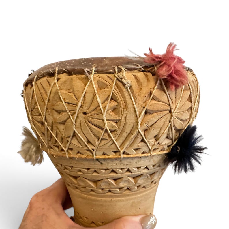May include: A hand-held wooden drum featuring intricate carved floral patterns on the exterior. The drum head is secured with a crisscross string lacing system. Decorative yarn pom-poms in shades of dusty pink, black, and cream are attached.