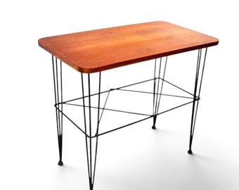 Peut inclure: Une table d'appoint rectangulaire avec un plateau en bois aux tons chauds et aux coins arrondis. La table est soutenue par une base en fil métallique noir qui comprend une étagère inférieure composée de fines tiges métalliques entrecroisées.