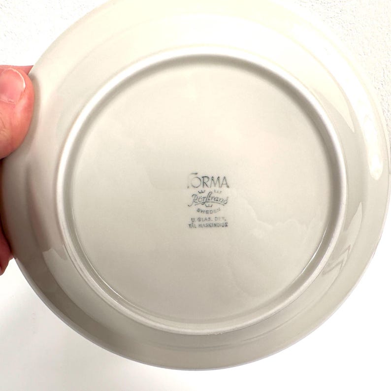May include: The bottom of a white ceramic plate with the text "FORMA  R&ouml;rstrand SWEDEN  U.GLAS.RET.  TIL MASKINVASK" printed in black.