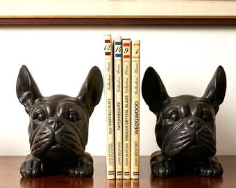 May include: A pair of black bulldog bookends flanking a row of books. The bookends are sculpted to resemble the dogs' faces. The books are titled "Old Sheffield Plate" and "Wedgwood". A vintage map is visible in the background.