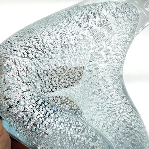 May include: A curved glass object featuring a clear, transparent outer layer and an inner layer filled with a dense, silver-colored metallic foil texture. The surface displays a complex, crackled, and bubbly appearance throughout the interior.
