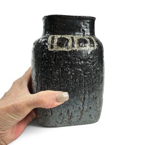 May include: A rectangular ceramic vase with a dark, textured black and grey finish. The surface features a rough, bark-like appearance with a prominent horizontal band near the top containing three distinct, light-colored rectangular motifs.