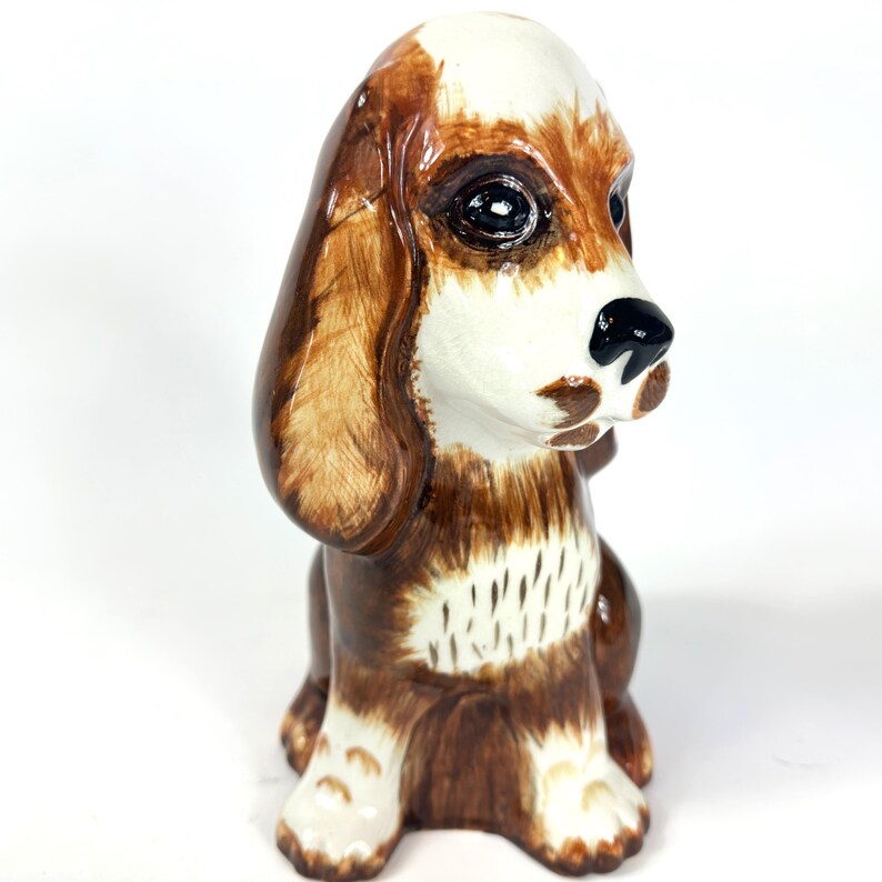 Peut inclure: Figurine de chien en c&eacute;ramique avec des marques marron et blanches. Le chien a de grands yeux expressifs, un nez noir et des oreilles tombantes. La figurine est assise, mettant en valeur ses d&eacute;tails et sa finition brillante.