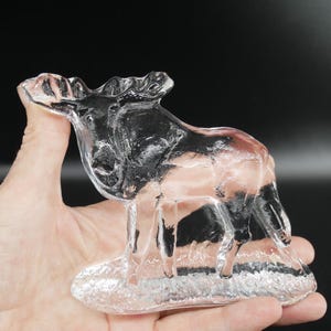 May include: A clear glass moose figurine. The moose is standing on a base, with detailed antlers and body features. The figurine is held in a hand against a dark background. The glass is transparent, allowing light to pass through.