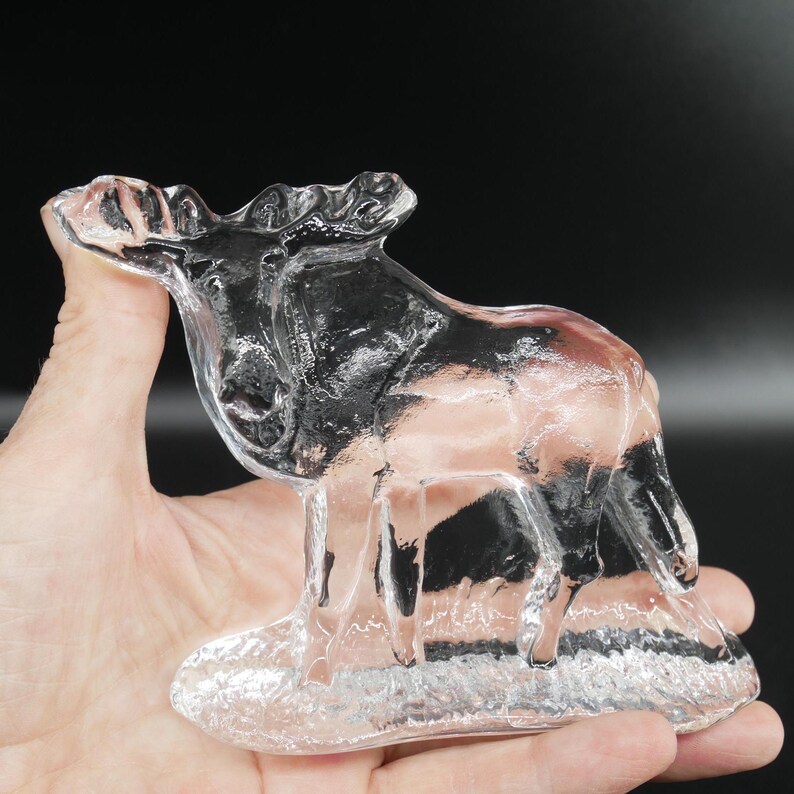 May include: A clear glass moose figurine. The moose is standing on a base, with detailed antlers and body features. The figurine is held in a hand against a dark background. The glass is transparent, allowing light to pass through.