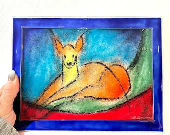 May include: A framed artwork featuring a stylized deer in shades of orange, yellow, and black, set against a backdrop of blue, green, and red. The artwork is encased in a vibrant blue frame.