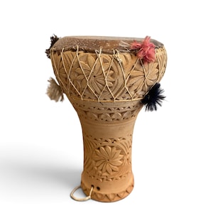 May include: A tan clay drum with a wide, rounded top and a narrow base. The exterior features intricate carved floral patterns and geometric designs. A dark skin head is secured with light-coloured cord lacing, accented by small pink, black, and white tufts.