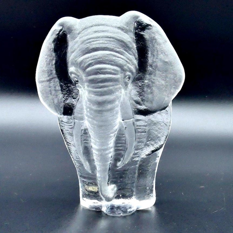 May include: A clear glass elephant ornament. The elephant is depicted standing with its trunk lowered and ears fanned out. The figurine has detailed wrinkles and folds, and the glass is transparent, allowing light to pass through.