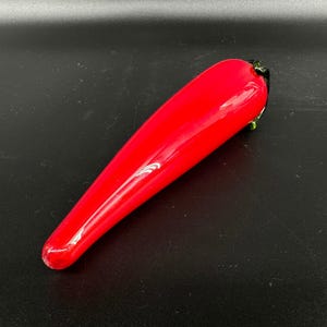 May include: A smooth, elongated glass sculpture shaped like a chili pepper. The body is a vibrant, glossy red, tapering to a rounded point at one end, while the opposite end features a small, dark green glass stem detail with a textured finish.