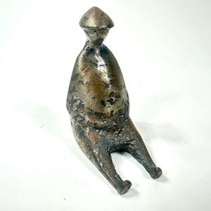 May include: A small, abstract bronze sculpture of a seated figure. The figure has a rounded, almost egg-shaped body and a pointed head. The sculpture has a rough, textured surface and a dark brown patina.