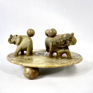 May include: A decorative stone elephant figurine set. The set includes three small elephant figurines and three spherical accents on a round, tiered base. The base is supported by a large spherical foot. The elephants and spheres are a light green color.