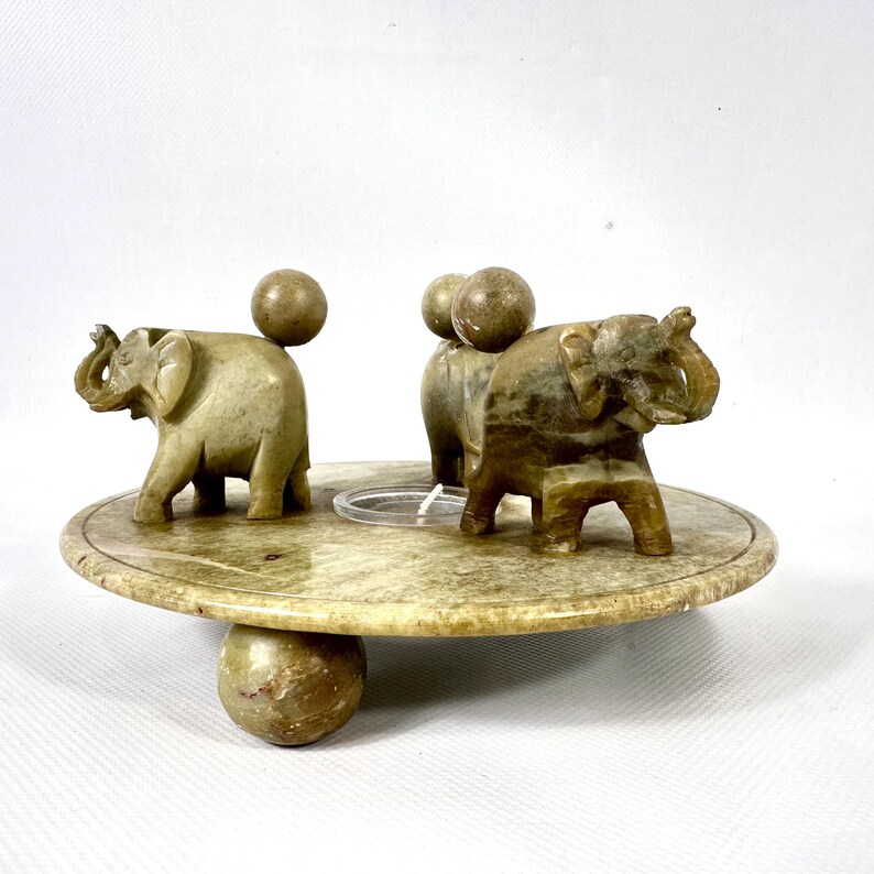 May include: A decorative stone elephant figurine set. The set includes three small elephant figurines and three spherical accents on a round, tiered base. The base is supported by a large spherical foot. The elephants and spheres are a light green color.