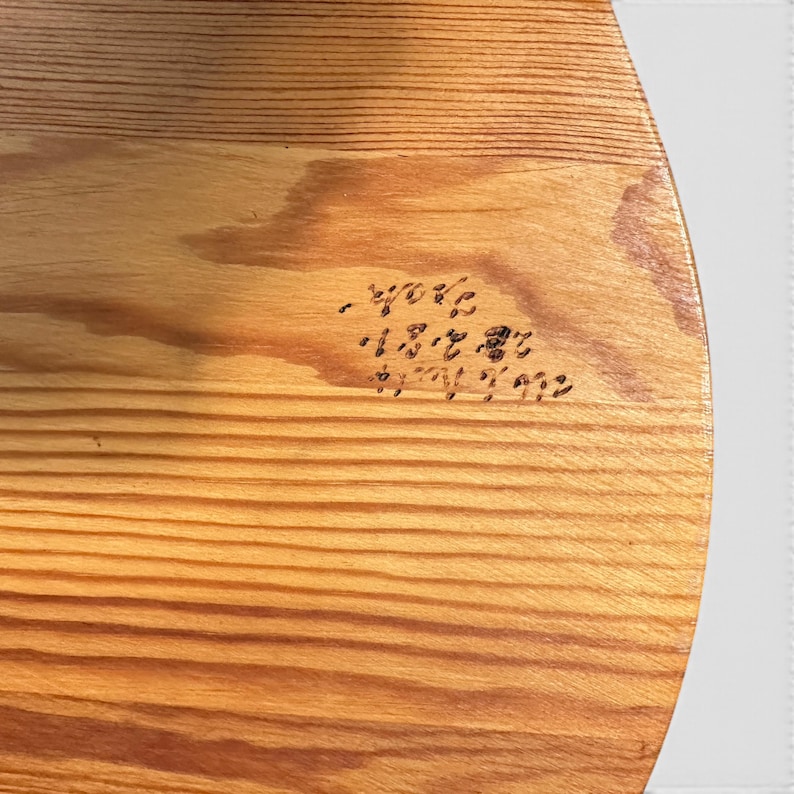 May include: A circular wooden board with a light brown natural grain pattern. The surface features a small, dark, hand-carved inscription in a cursive script that appears to be a signature or date, positioned slightly off-centre on the wood.