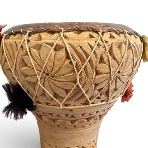 May include: A hand drum with a light brown wooden body featuring carved floral patterns and a dark brown skin head. The drum is secured with a crisscross pattern of light-coloured cord and decorated with small black and orange tufts of fibre.