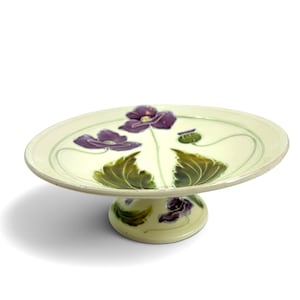 Könnte beinhalten: Eine Keramikschale auf einem Fuß mit cremefarbener Glasur, verziert mit handgemalten violetten Mohnblumen, grünen Blättern und dünnen grünen Stielen. Die Schale hat ein breites, flaches rundes Becken auf einem stabilen, passenden Fuß.