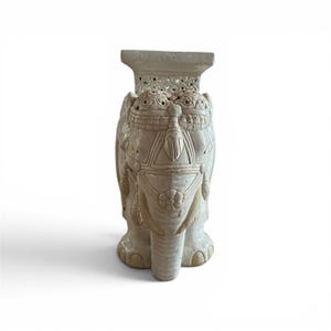 Może przedstawiać: Kremowy ceramiczny stołek ogrodowy w kształcie słonia z misternymi rzeźbionymi detalami, płaskim prostokątnym blatem i ozdobnymi motywami siodła. Słoń stoi w pozycji skierowanej do przodu z trąbą skierowaną w d&oacute;ł, prezentując błyszczące wykończenie.