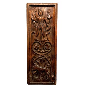 May include: A rectangular dark brown wooden wall panel featuring intricate hand-carved relief designs. The top section depicts a hooded figure holding a tool, while the lower section shows a four-legged animal amidst stylized foliage and circular motifs.