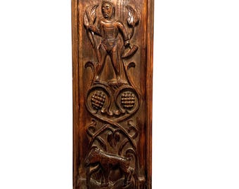 May include: A rectangular dark brown wooden wall panel featuring intricate hand-carved relief designs. The top section depicts a hooded figure holding a tool, while the lower section shows a four-legged animal amidst stylized foliage and circular motifs.
