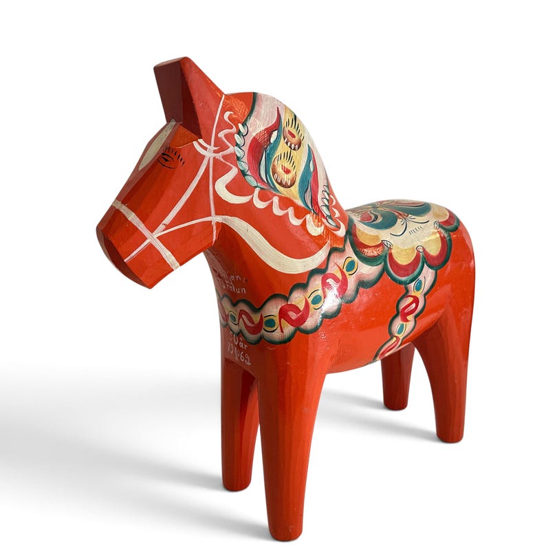 Puede incluir: Una figura de caballo Dala de madera en color naranja brillante con patrones florales y de volutas pintados a mano en blanco, azul y verde. El costado del caballo muestra un texto en blanco que dice 50 &aring;r y 25/6 62, indicando una fecha.