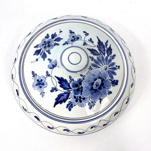 May include: A white ceramic lid decorated with blue floral patterns. The lid has a central knob and a patterned border. The floral design includes various sizes of flowers and leaves, all in shades of blue.
