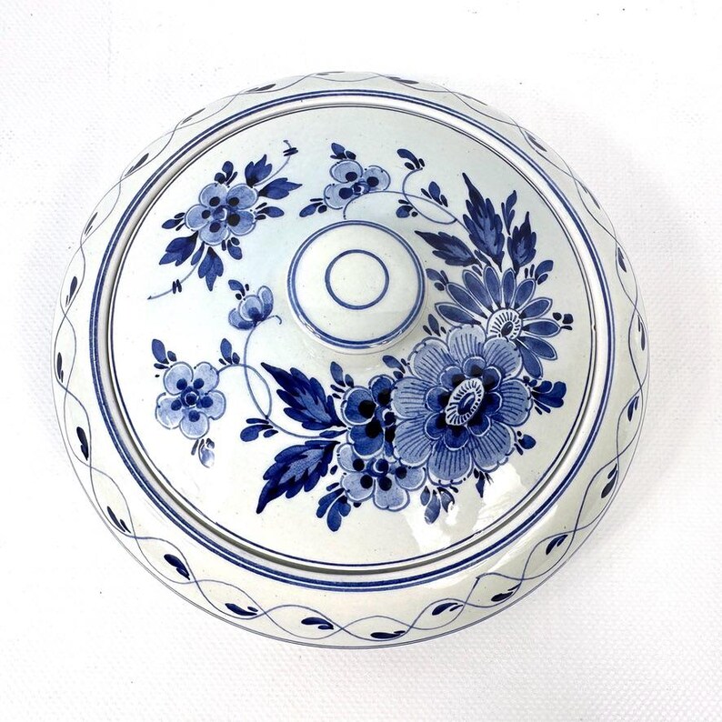May include: A white ceramic lid decorated with blue floral patterns. The lid has a central knob and a patterned border. The floral design includes various sizes of flowers and leaves, all in shades of blue.