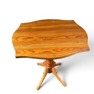 May include: A wooden pedestal table featuring a light brown finish with a visible wood grain pattern. The tabletop has a distinct scalloped edge design, supported by a central column base that branches into three angled legs.