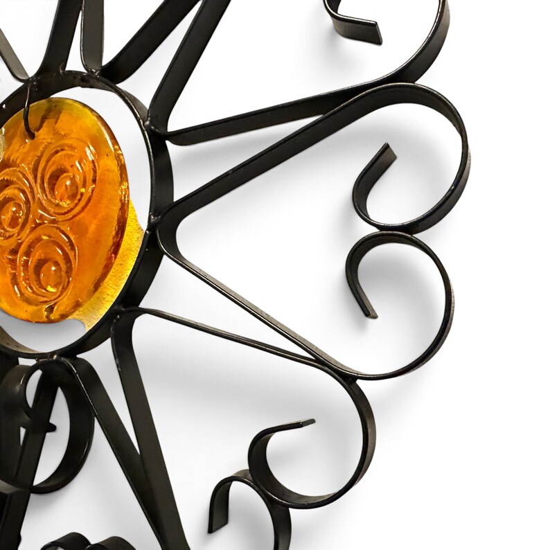 Peut inclure: Une d&eacute;coration murale compos&eacute;e d'un cadre en m&eacute;tal noir avec des p&eacute;tales incurv&eacute;s en forme de volutes dispos&eacute;s en cercle. Au centre, un insert en verre ambr&eacute; affiche trois cercles concentriques en relief sur sa surface translucide.