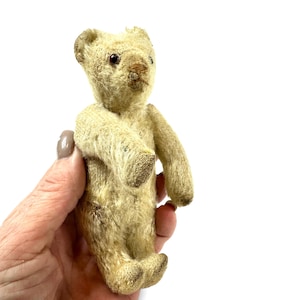 May include: A small, vintage-style teddy bear with worn, cream-colored fur, dark brown bead eyes, and a stitched nose. The bear features articulated limbs, rounded ears, and a textured, aged appearance throughout its entire body structure.